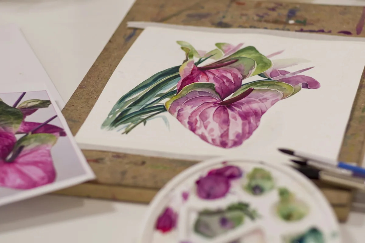 Lámina de acuarela botánica en curso sobre la mesa de trabajo, con paleta de colores y pinceles al lado